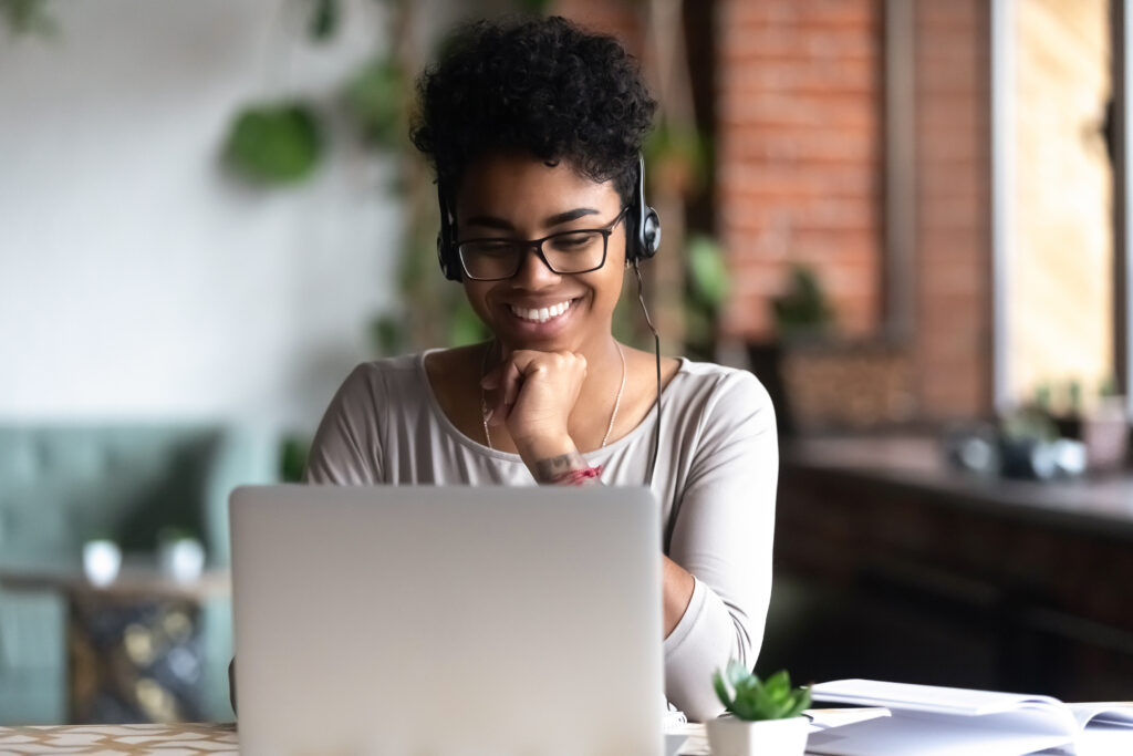 Smiling woman wearing headphones, engaged with a laptop in a cozy workspace, representing online learning and productivity.